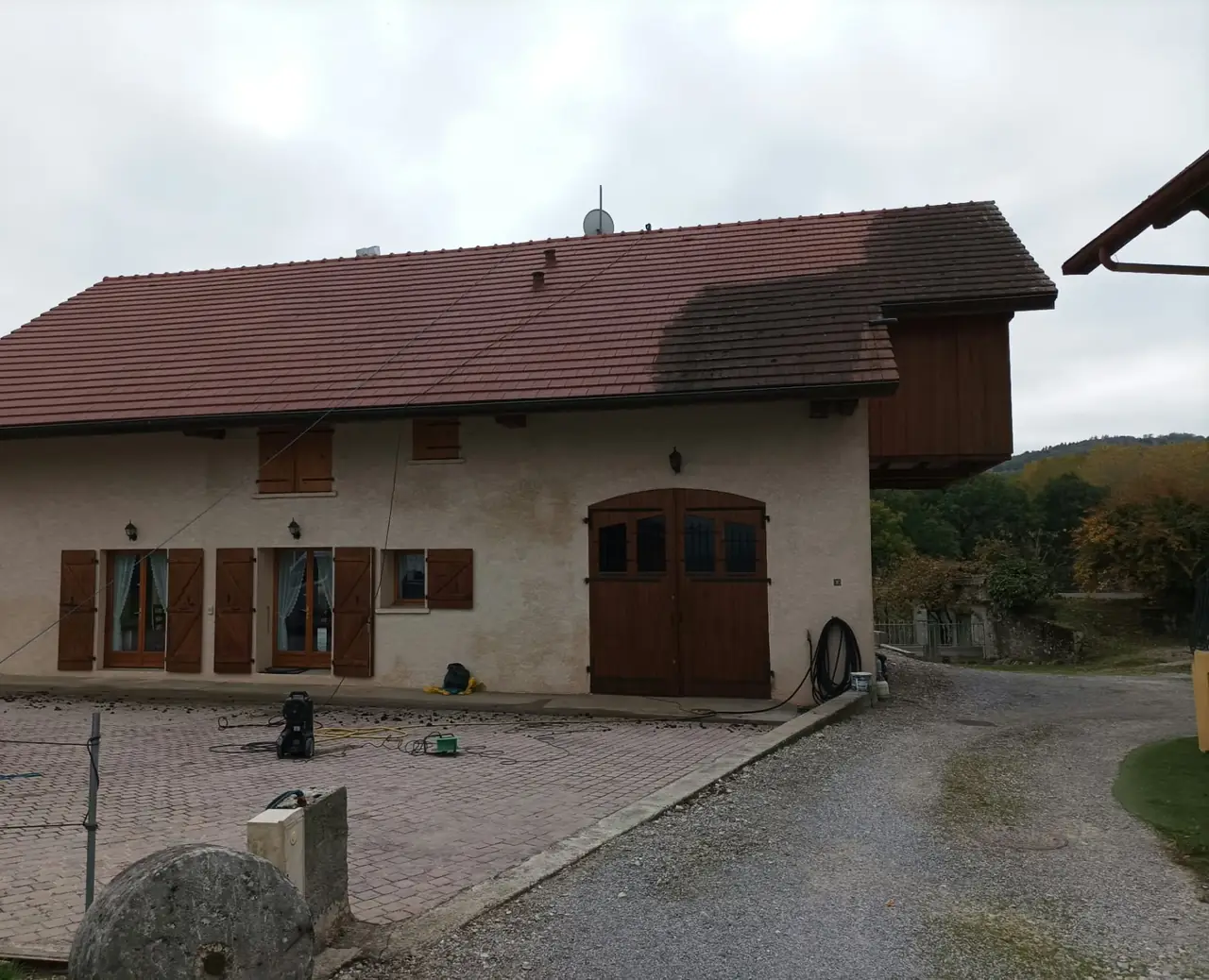 Maison aux volets en bois dont la toiture est à moitié nettoyée : tuiles rouges propres à gauche, encore sombres à droite ; nettoyeur haute pression dans la cour pavée